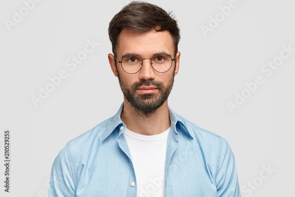 Fototapeta Headshot of attractive bearded young European male with serious confident expression, looks directly at camera, wears spectacles, isolated over white background, thinks about starting family life.