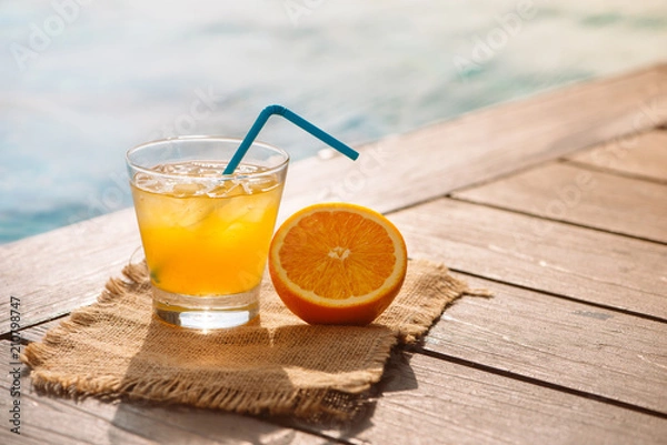Fototapeta Close up of screwdriver cocktail alcohol drink with orange juice, slices and ice standing near the pool. Refreshing iced lemonade beverage in glass by the poolside. Sun glares. Background, copy space.