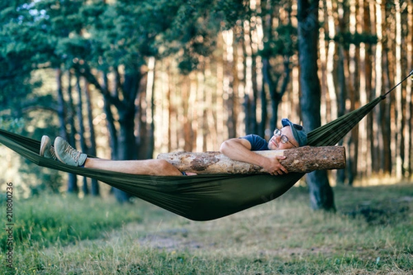 Obraz Odd weird strange unusual male person. Outlandish funny crazy foolish man sleeping in hammock with huge wooden log at nature among trees.  Wood and forest lover. With beam in bed.  Leisure lifestyle.