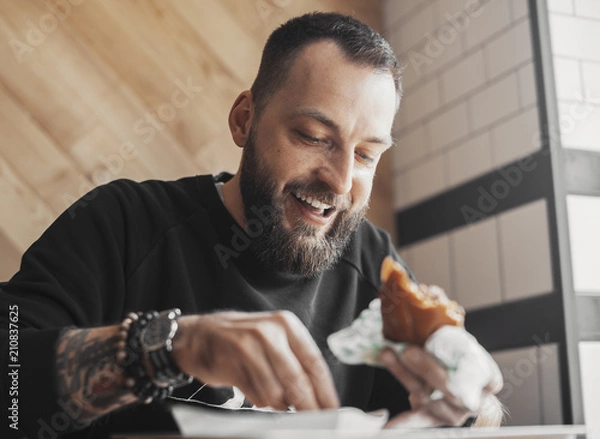 Obraz Young bearded man eating burger and smiling close up.