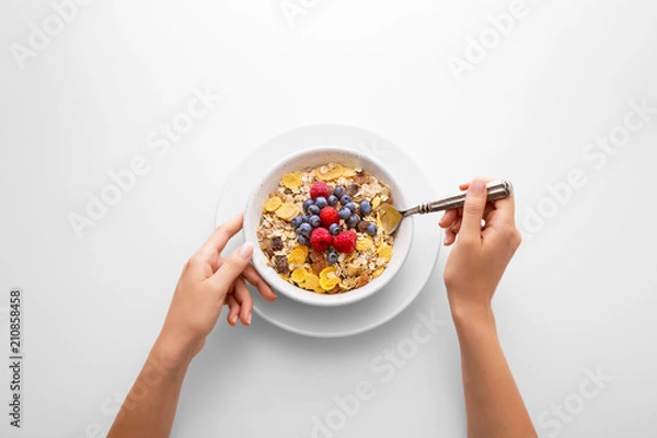 Fototapeta Young woman eating muesli cereal breakfast with berry fruit in a white bowl viewed directly from above. Female hand holding a spoon with fresh rich in fiber granola dish viewed from above. Top view.