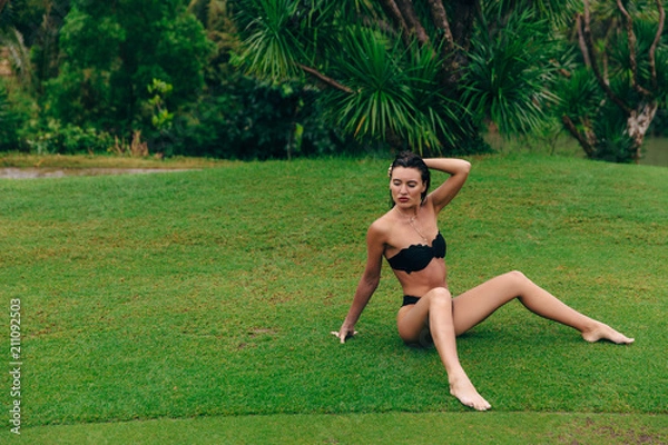 Fototapeta A dark-haired dark-haired girl with red lips in a black swimsuit without a britewalk uninhibited sits on the green grass, straightens long hair, enjoys cloudy weather.