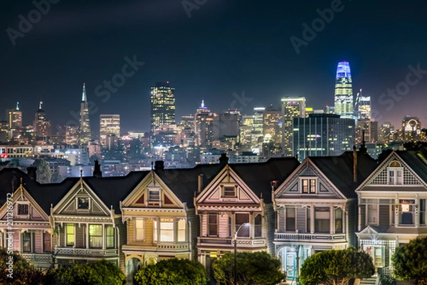 Fototapeta Oparte na nocnej panoramie miasta San Francisco w Kalifornii, domy z epoki wiktoriańskiej w pobliżu parku Alamo Square są pomalowane na kolory, aby podkreślić ich detale architektoniczne.