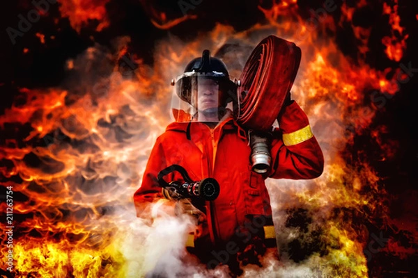 Fototapeta fireman wearing firefighter suit holding a fire hose and nozzle to using water and extinguisher to fighting with fire flame in an emergency situation., burning building in background.