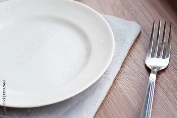 Obraz Empty white plate on a gray napkin with fork and glass on brown table. Table setting, preparation for meals.