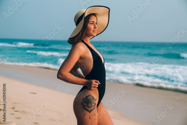 Fototapeta Side view of a young pretty girl in a black swimsuit, has a sexy tattoo on her thigh, wears a fashionable hat, looks at the sun, walks along the beach.