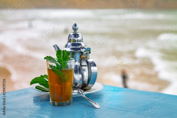 Fototapeta A glass of mint tea with an iron kettle, a sprig of mint and a large slice of sugar. A traditional Berber drink mint tea in the background of the Atlantic Ocean and mountains. Africa, Morocco, Agadir