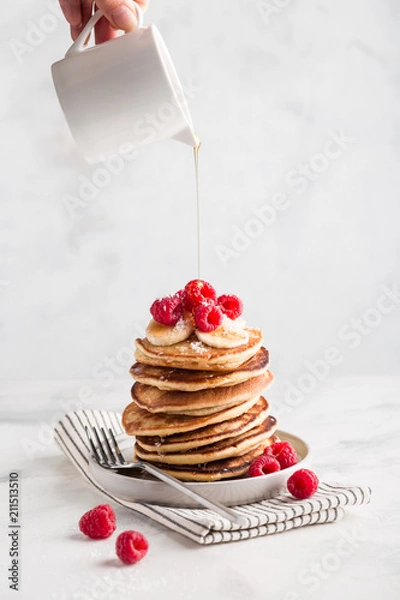 Fototapeta Hand pouring maple syrup on stack of homemade pancakes with fresh raspberries on light marble background