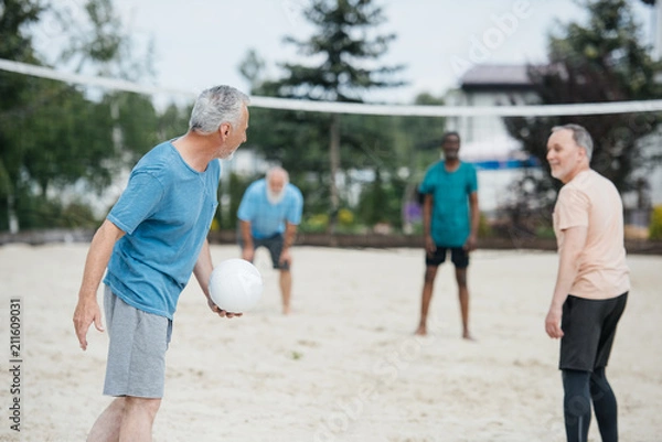 Fototapeta selective focus of multicultural old friends playing volleyball on beach on summer day
