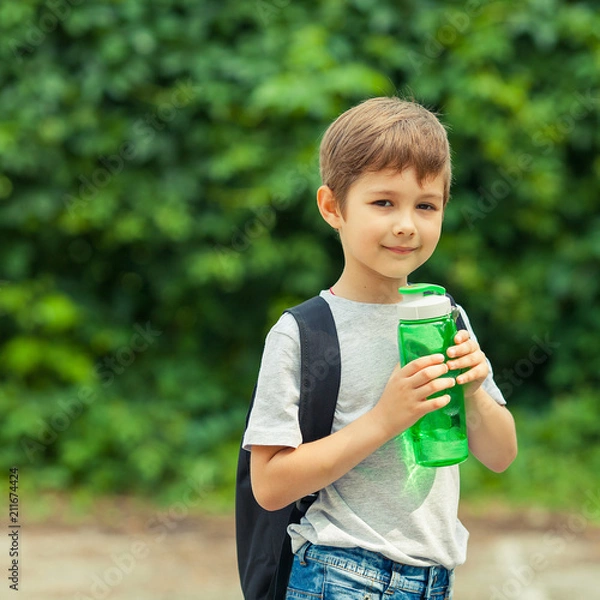 Fototapeta Little boy drinking water from a bottle, hot summer. Child is traveling with a backpack. Vacation in the woods.