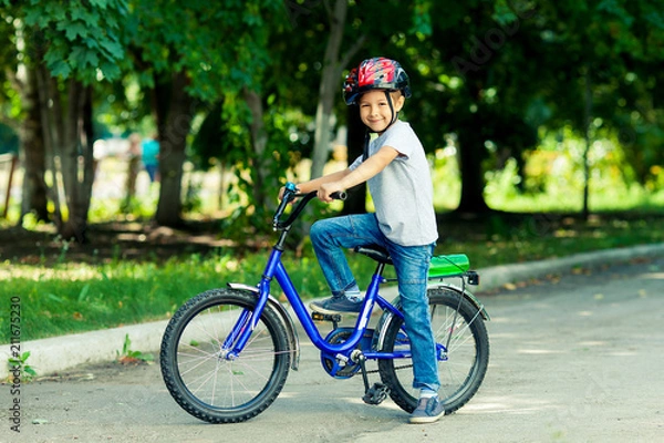 Fototapeta Little boy learns to ride a bike in the park near the home. Portrait of a cute kid on bicycle. Happy smiling child in helmet riding a cycling.