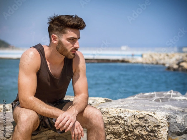 Obraz Attractive fit athletic young man soaking in the sun on seaside boardwalk or seafront, sitting on rock, wearing black tank-top