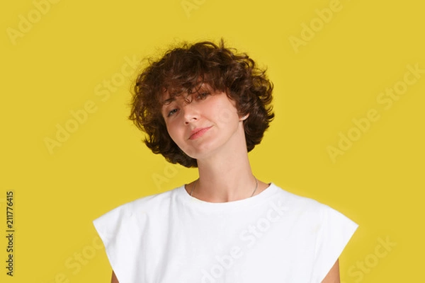 Fototapeta Portrait of pleased beautiful European female with curly brunette hair, dressed in casual white t shirt on yellow background. Attractive woman student has confident look. Facial expressions concept