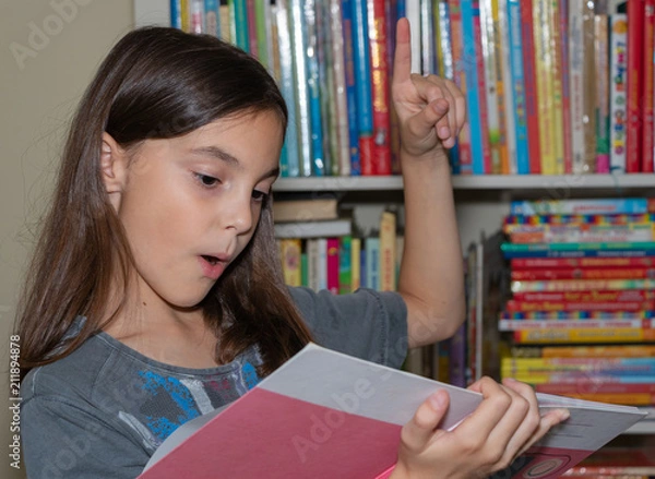 Fototapeta background, cabinet with colored books. a beautiful girl in her arms has a light pink book. reads, looks and is surprised.