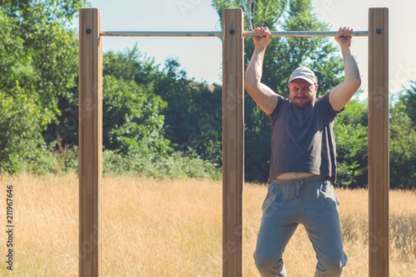 Fototapeta Young man with excess weight doing pull up exercise on horizontal bar