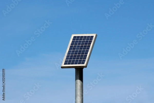 Fototapeta Small solar panel mounted high on top of thick grey metal pole on warm sunny winter day with clear blue sky in background
