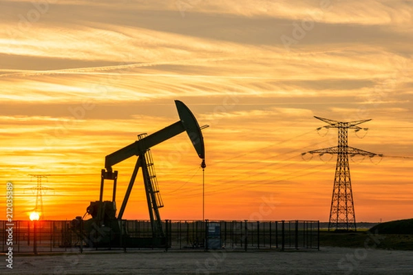 Fototapeta View of a pumpjack at sunset pumping oil out of a well with the silhouettes of transmission towers supporting an overhead power line in the background against a red sky.