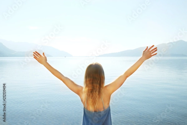 Fototapeta Happy young woman with hands up on sandy beach watching islands in the sea on sunny day. Hipster female in loose denim dress on ocean shore having fun watching waves, sunbathing. Background, close up