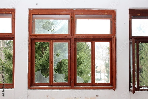 Fototapeta Renovated wooden window frame with broken glass and tall pine trees in background surrounded with dilapidated wall