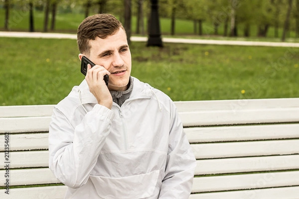 Obraz Молодой парень европейской внешности с серьезным лицом сидит на лавочке говорит по телефону