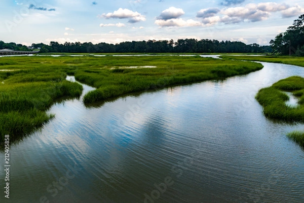 Fototapeta Wetlands river runs through reeds and into a coastal river underneath a dark and brooding sky