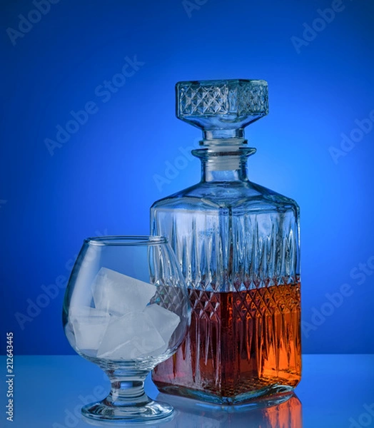Fototapeta Glass of brandy with ice and a square decanter isolated on a blue background