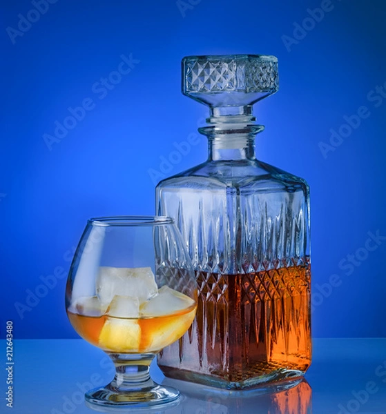 Fototapeta Glass of brandy with ice and a square decanter isolated on a blue background