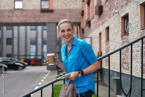 Fototapeta Young girl with tablet and coffee stands on a city street. She holds a backpack in her hands and laughs.