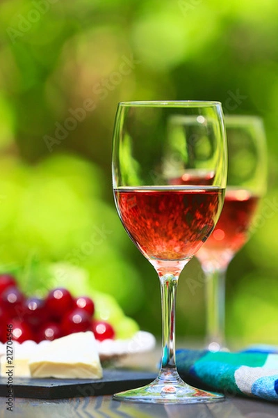 Fototapeta Two glasses of Shinning Rose Wine on wooden table with fresh Grapes and Cheese, bright and green outdoors background 2