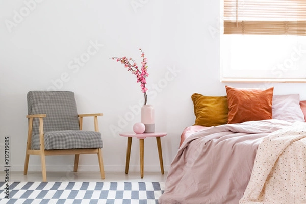 Fototapeta Flowers on table between patterned armchair and pink bed in bedroom interior with window. Real photo