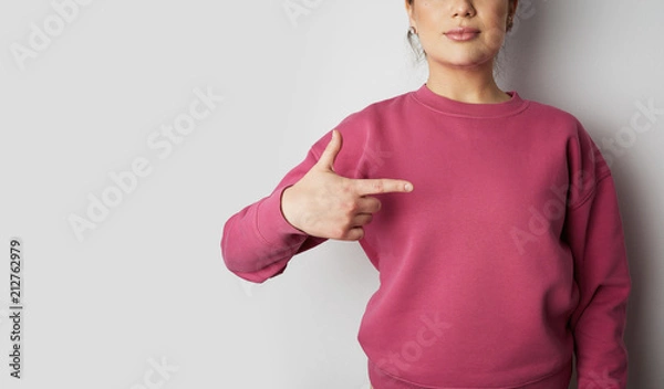 Fototapeta Young hipster girl wearing pink hoody and pointing finger with space for your logo or design. Mock-up of pink hoody on white empty wall in the background. Copy paste text space