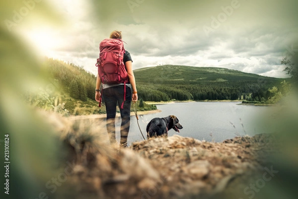 Fototapeta young woman with backpack and german shepherd dog puppy standing on mountain in front of forest and lake