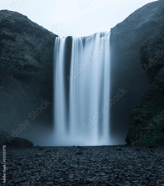 Fototapeta Beautiful Waterfall in Iceland from the front with rough looking water falling off the hill in a dark look with black stones in the foreground and no people in front during night