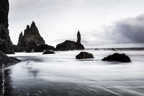 Fototapeta Rocks on a black sand beach in Iceland with reflection in the Sea and a dark sky in misty moody weather with dark colors and a rough sea in landscape format with a storm approaching