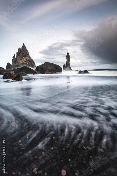 Fototapeta Rocks on a black sand beach in Iceland with reflection in the Sea and a dark sky in misty moody weather with dark colors and a rough sea in landscape format with a storm approaching