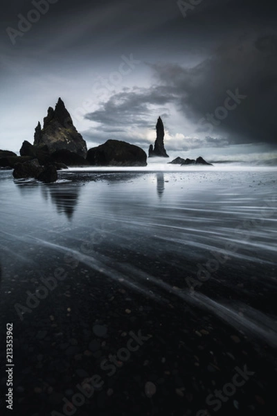 Fototapeta Rocks on a black sand beach in Iceland with reflection in the Sea and a dark sky in misty moody weather with dark colors and a rough sea in landscape format with a storm approaching