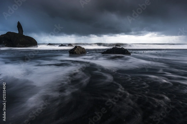 Fototapeta Rocks on a black sand beach in Iceland with reflection in the Sea and a dark sky in misty moody weather with dark colors and a rough sea in landscape format with a storm approaching