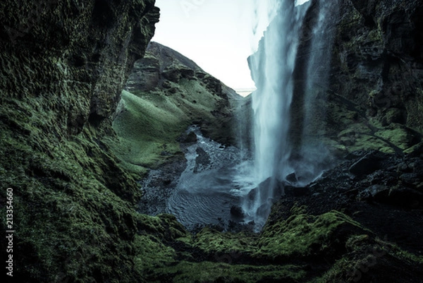 Fototapeta Wild waterfall in a green cave with green moos and wet black sand and stones with a creek flowing through a green grass covered rocky hilly valley surrounded by hills in Iceland