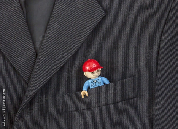 Fototapeta in the breast pocket of a dark black man's jacket in a light strip is a toy from the designer of a little man in a red cap