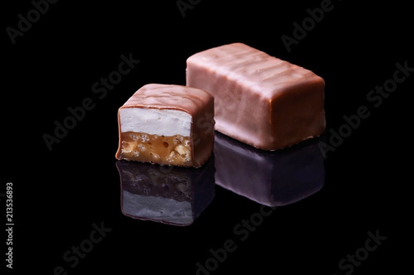 Fototapeta Chocolate bar in a cut on a black specular background. Isolated chocolate dessert with caramel filling and nougat. Smooth cut of the chocolate.