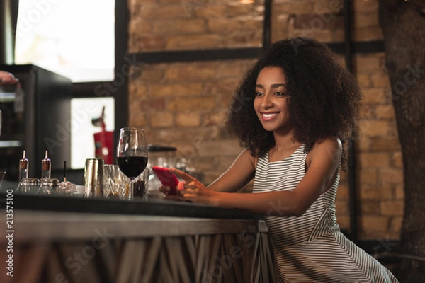 Fototapeta Good news. Petite curly woman sitting at the bar counter and smiling happily having received a message with good news on her smartphone
