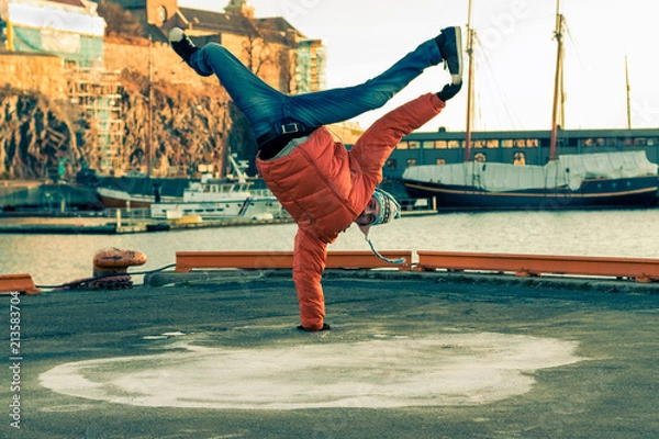 Obraz Young guy dancing break dance on the waterfront in an orange jacket in winter.