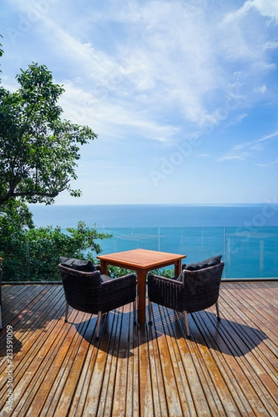 Obraz Table and rattan outdoor armchair setting on wooden deck and glass balcony with panoramic Andaman ocean sea view, green tree and blue sky background on sunny day