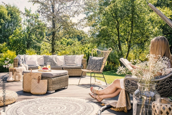 Fototapeta Kobieta czytająca książkę relaksując się na tarasie z meblami rattanowymi w ogrodzie. Prawdziwe zdjęcie