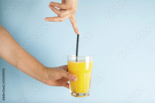 Fototapeta Hand holding glass of orange juice black straw isolated on blue background