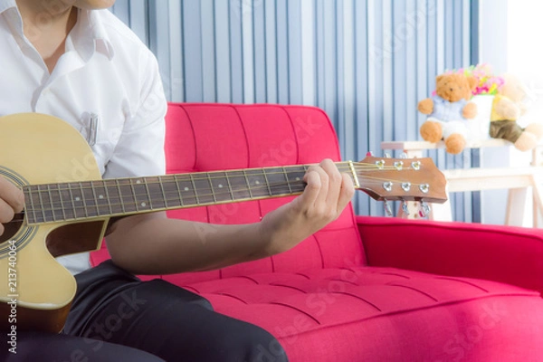 Fototapeta A man is playing music by classic guitar on vivid red long chair with soft yellow light in living room