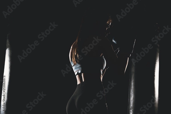 Fototapeta Woman with black boxing wraps and boxing gloves on hands boxing in dark with punching bag. Boxer girl silhouette