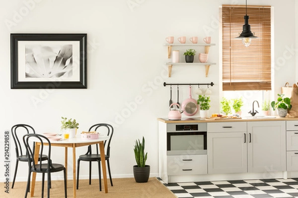 Fototapeta Painting in a black frame in a dining room interior with a table and chairs next to a kitchen interior with tools, plants and cupboards, Real photo