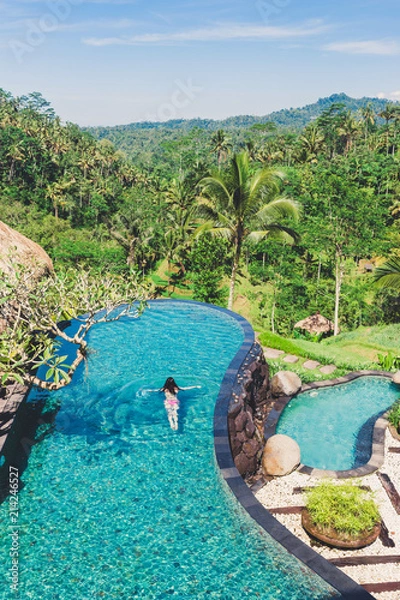 Fototapeta Girl is swimming in a large beautiful pool against the backdrop of lush tropical vegetation. A young woman swims in an outdoor pool with a beautiful view of palms, Bali, Indonesia. View from above.