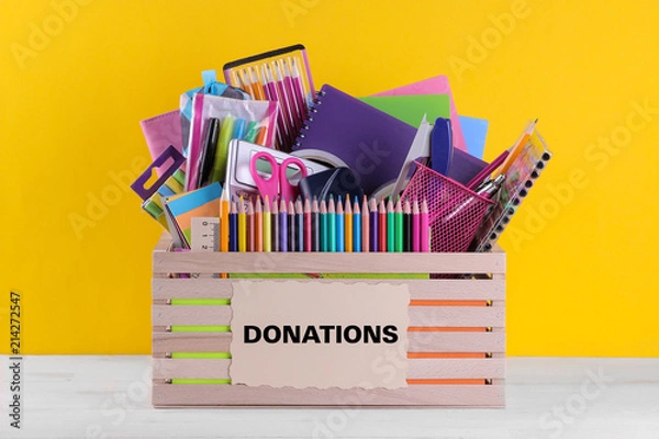 Obraz Box with various school and office supplies with a sign on a bright yellow background. Donation concept